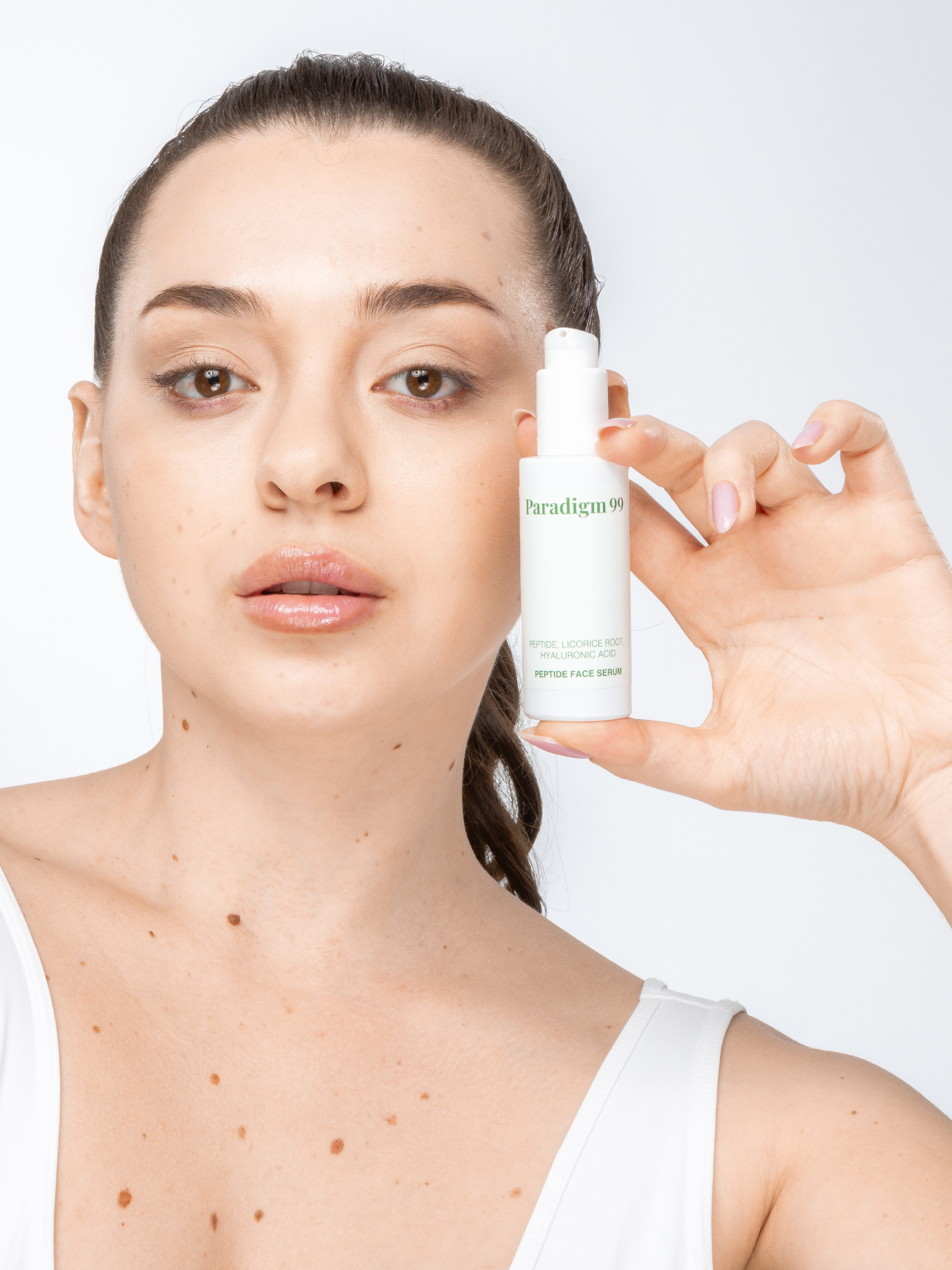 A woman with brown hair in a ponytail holding a paradigm99 peptide serum in a light green glass bottle with a dispenser. She has beautiful, well cared for skin with a few moles on her right cheek.