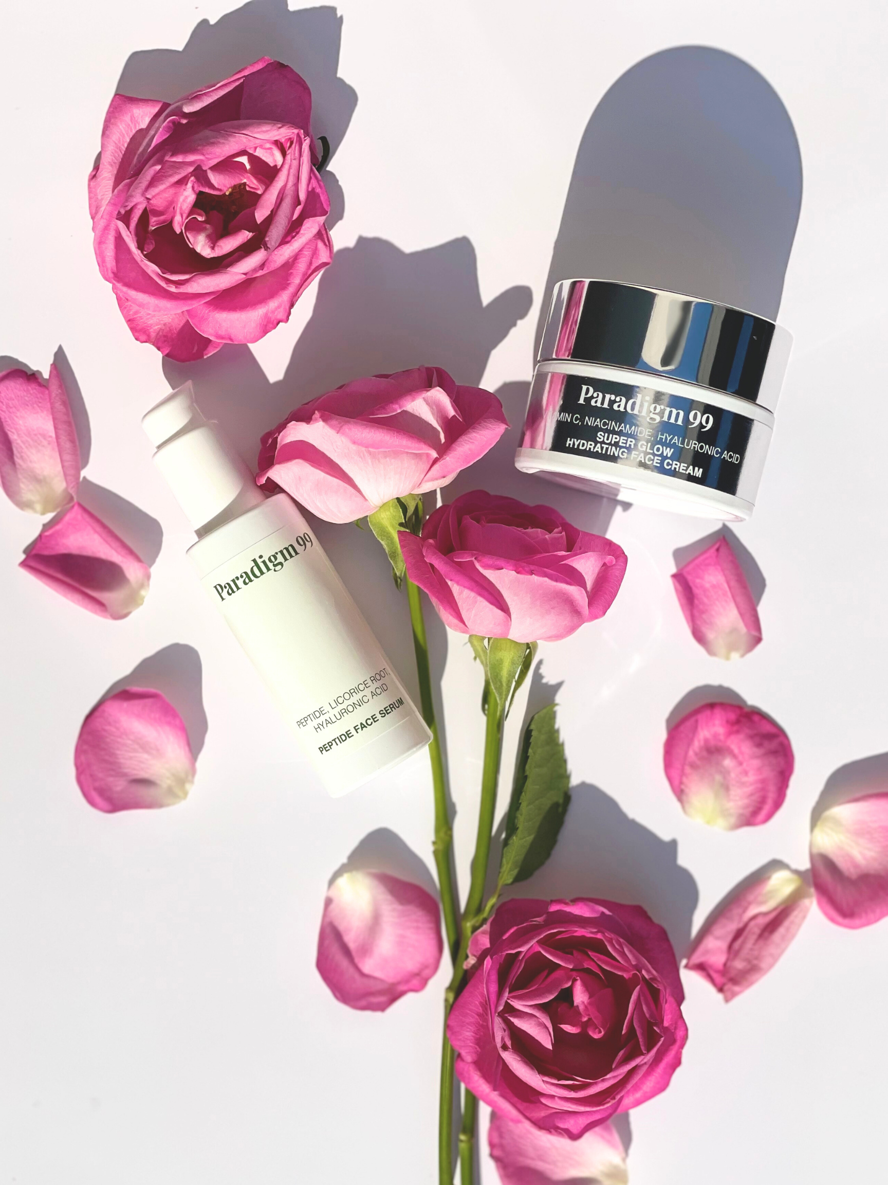 Two skincare bottles labeled 'Paradigm 99' surrounded by pink roses on a light background. First product is Peptide Face Serum with Peptide, Licorice Root, Hyaluronic Acid. Second product Super Glow Hydrating Cream with Vitamin C, Niacinamide, Hyaluronic Acid. 