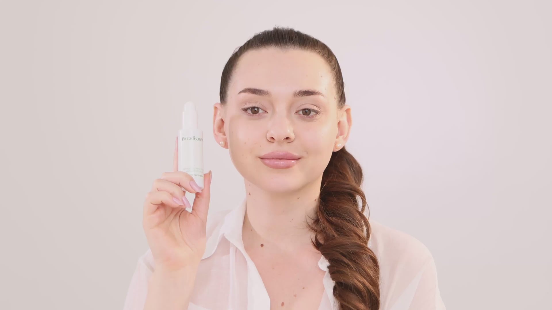 Video of a woman applying peptide serum Paradigm 99, pressing the pump to dispense a milky-colored serum. She spreads it over her forehead, cheeks, and nose, admiring her nourished and refreshed skin after skincare.
